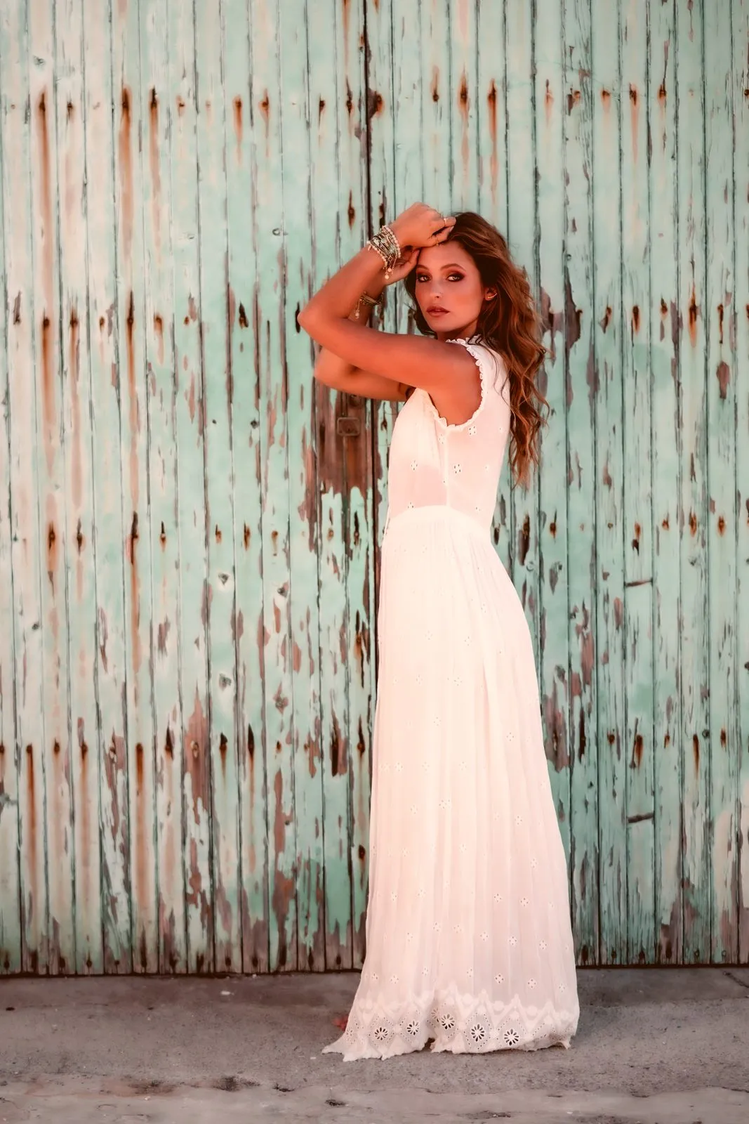 A model in a long white lace dress before a peeling turquoise door, a hand in her hair, Dreamers Ibiza