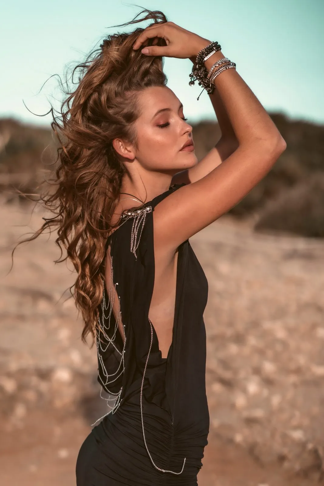 A model in black with a chain back-necklace, hands in her hair on the beach, Dreamers Ibiza