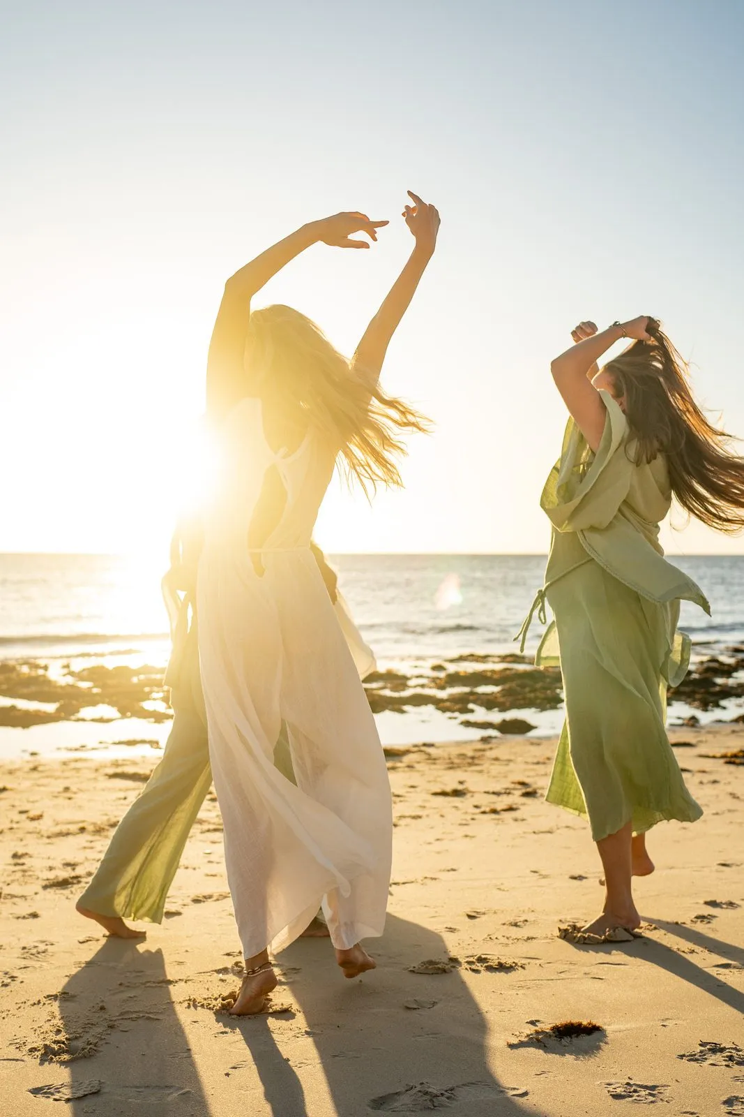 Two women in flowing dresses dancing on the beach at sunset, backlit by the sun, Nomadica
