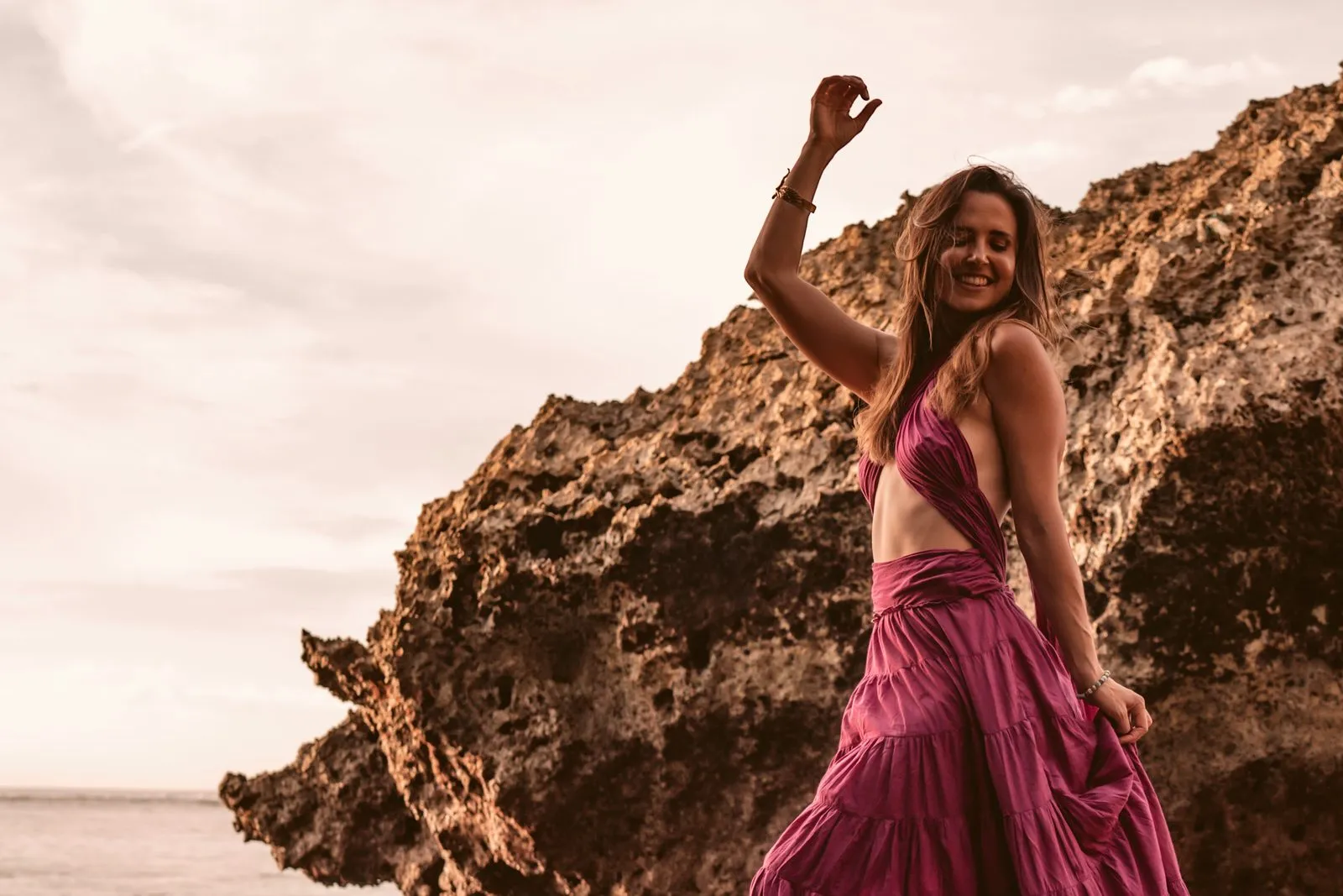A woman in a plum dress twirling at the foot of the limestone cliffs at Uluwatu, Bali