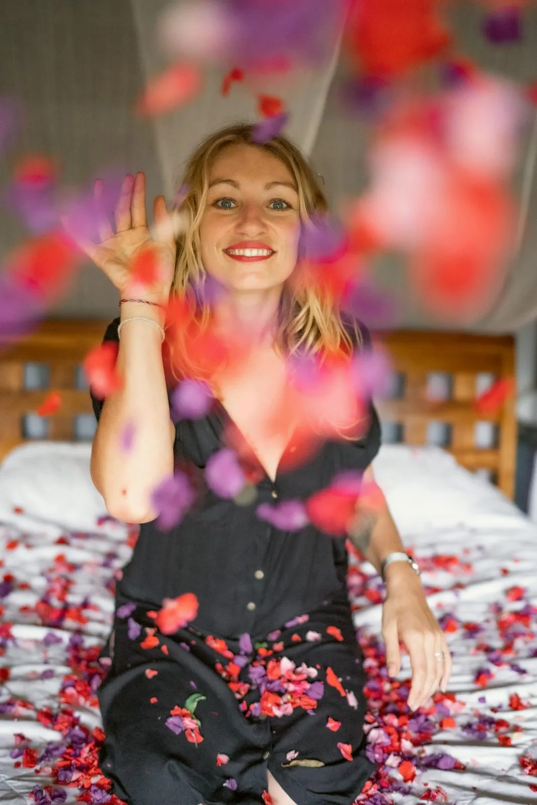 A smiling woman on a bed tossing rose petals into the air, petals falling all around her