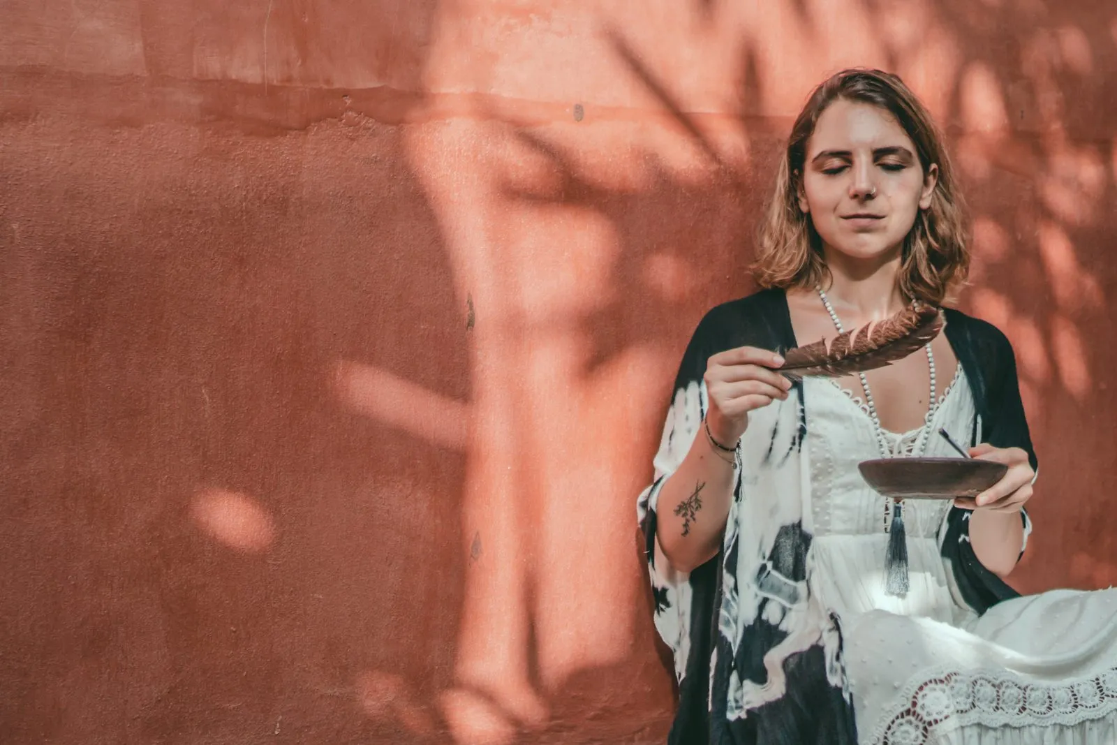 A woman holding a bowl of smoking sage against a terracotta wall, in the dappled shadow of leaves