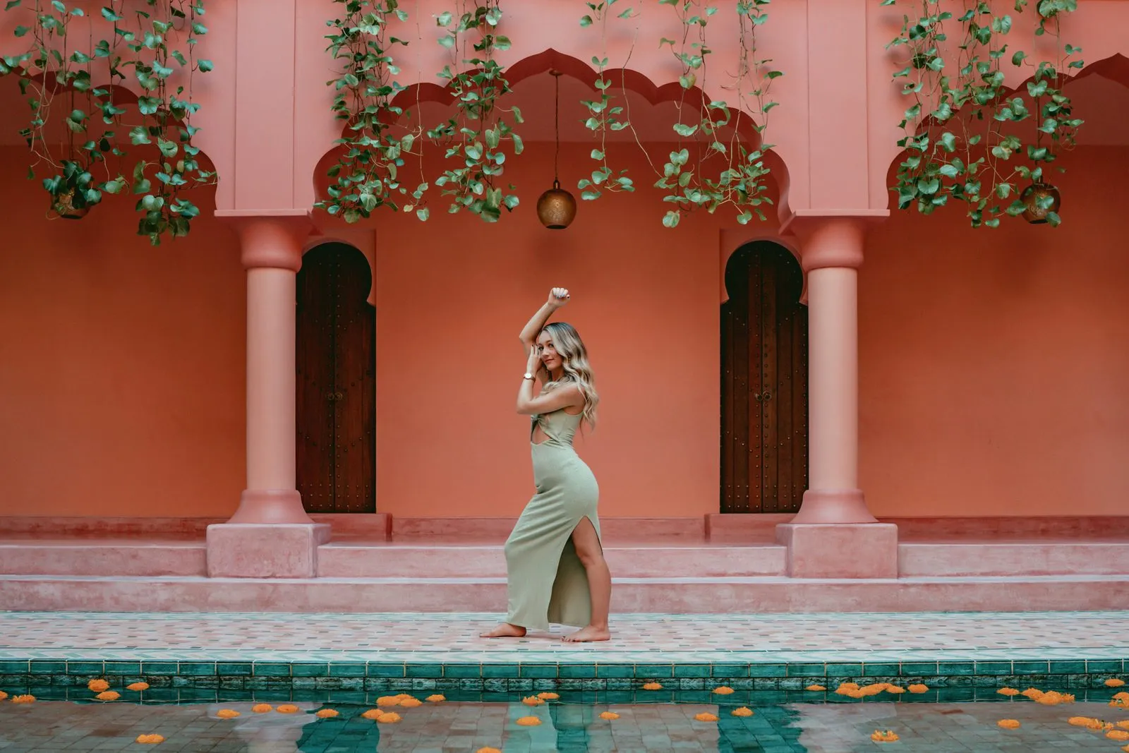 A woman in green at the edge of a pool in a pink courtyard, marigolds floating on the water and vines over the arches