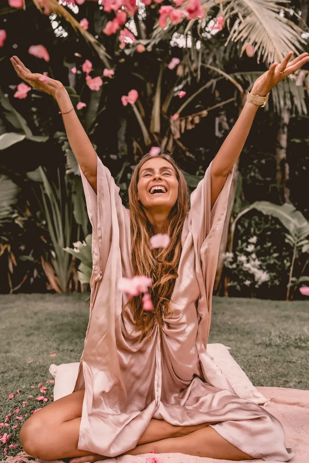 A woman in a silk robe kneeling on the grass with her arms flung up, pink petals falling from the frangipani above