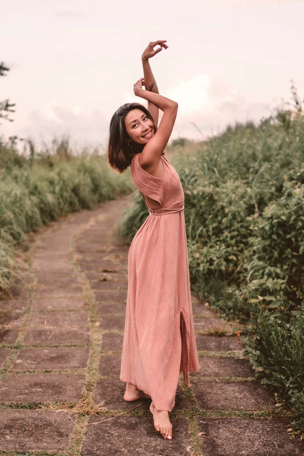 A woman in a dusty pink dress on a stone path between rice fields, one arm raised over her head