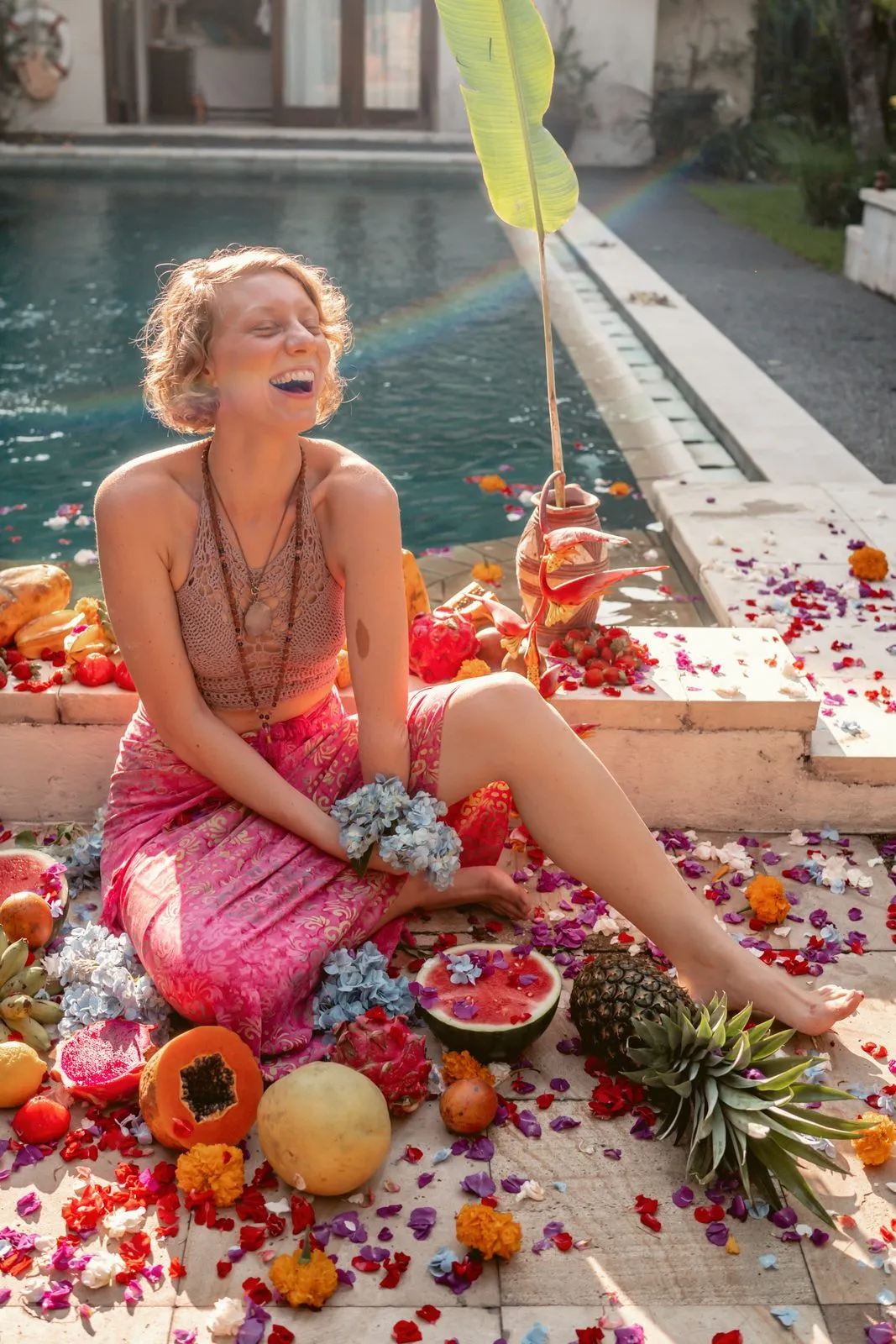 A laughing woman in pink sitting by a pool among papaya, pineapple, pomegranate and scattered flowers
