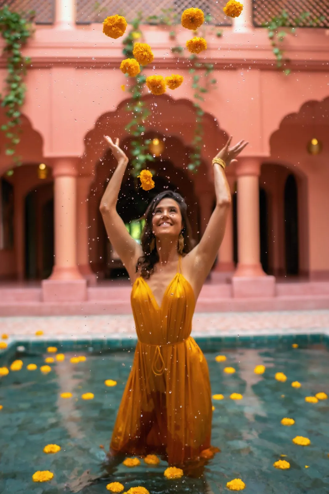 A woman in a yellow dress waist deep in a pool, throwing marigolds into the air in a pink courtyard