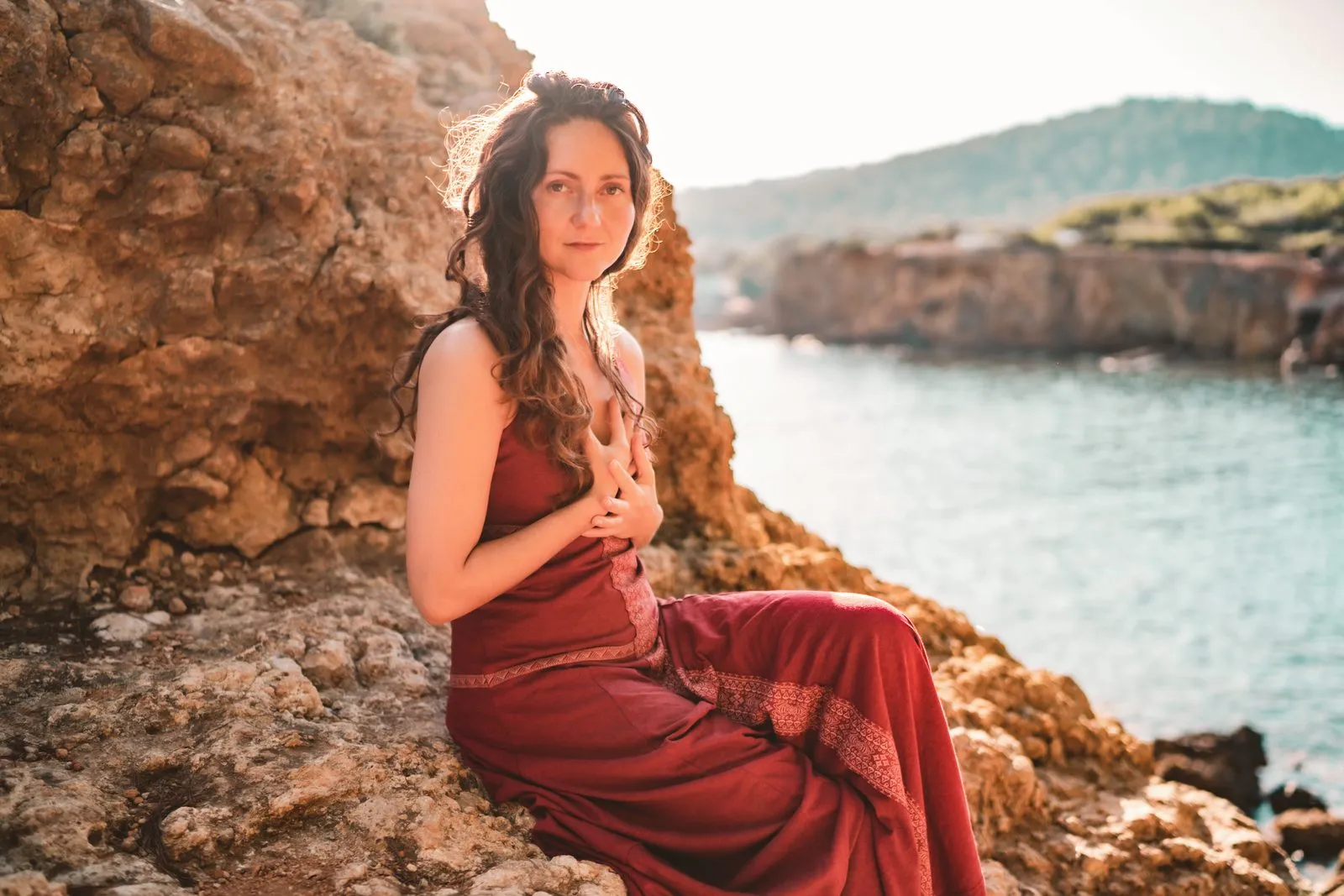 A woman in a red dress sitting on the rocks above a cove, the cliffs and the sea behind her
