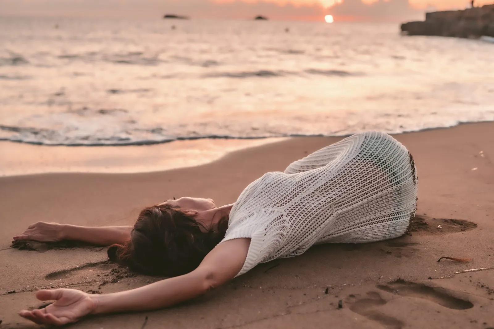A woman in a crochet dress lying back on the wet sand at the edge of the sea at sunset