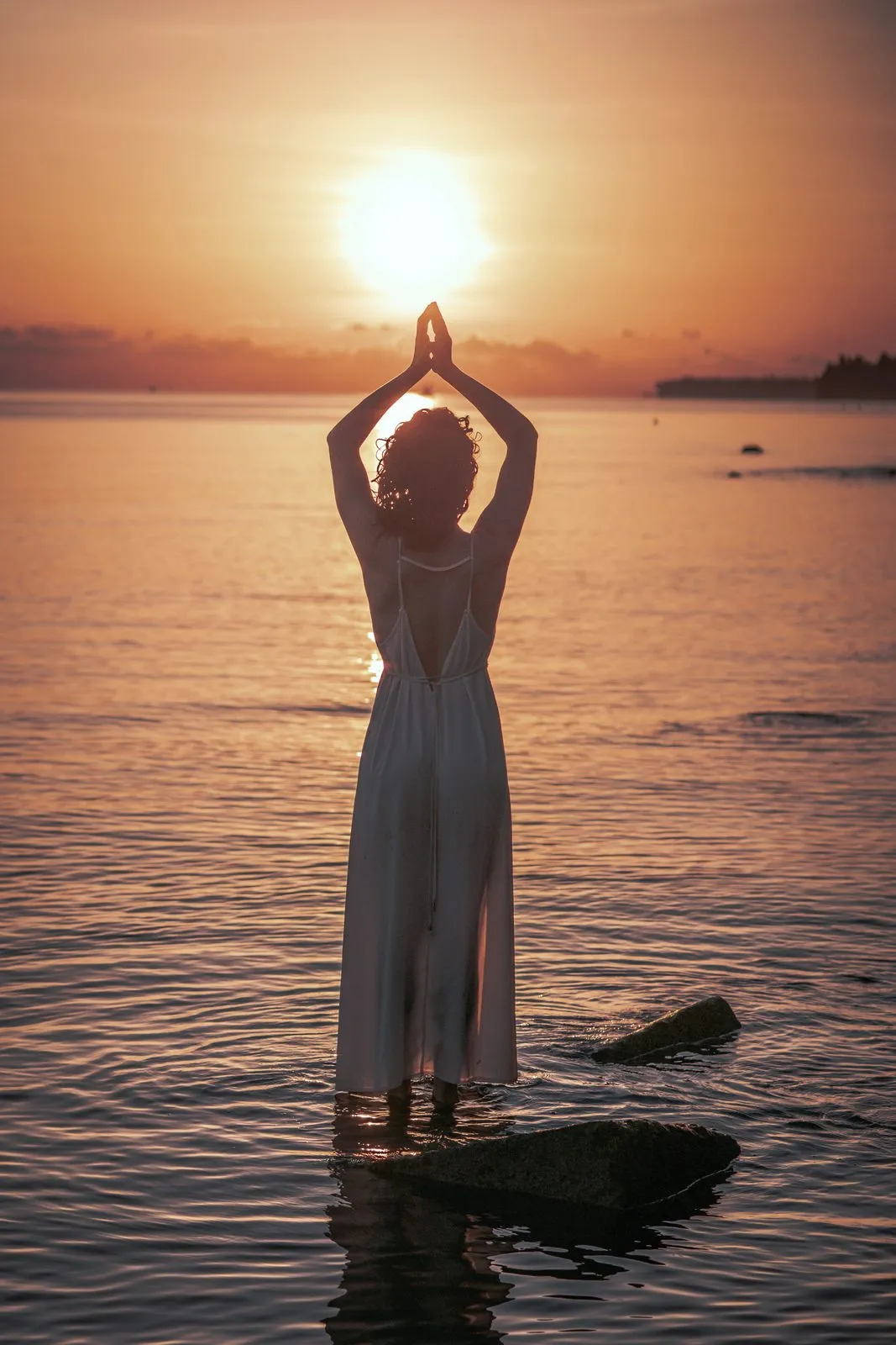 A woman standing in the shallows at sunset, her hands joined above her head around the sun