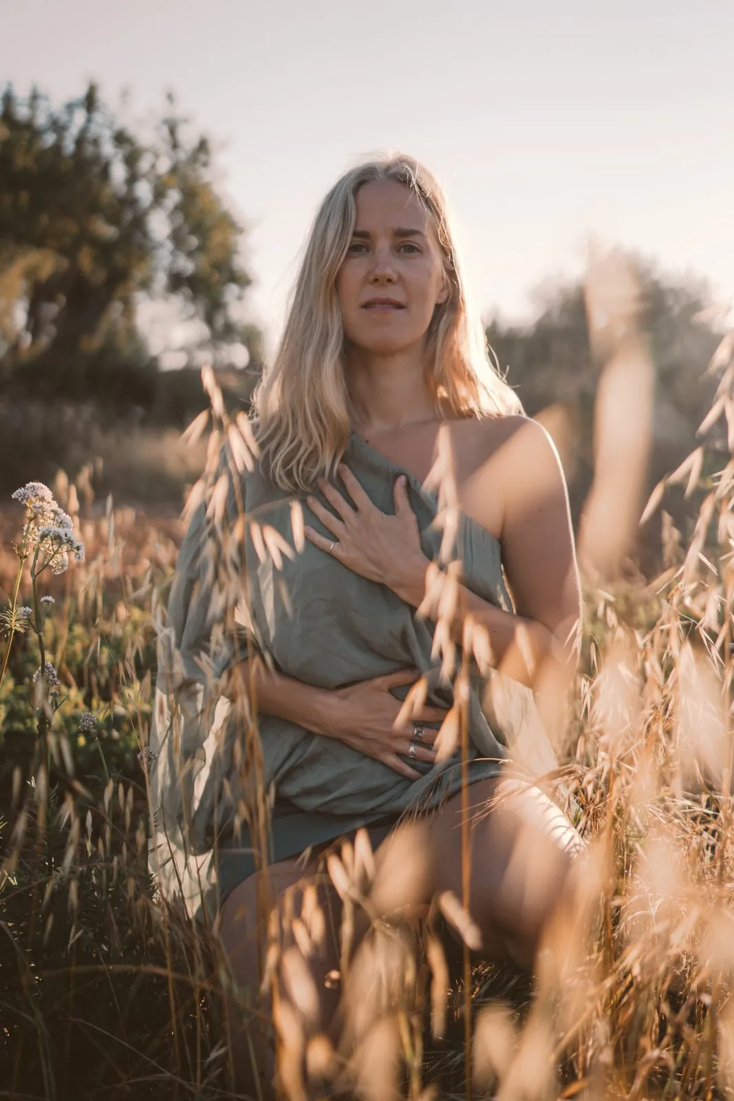 A woman in grey standing in long golden grass with a hand on her heart, lit from behind