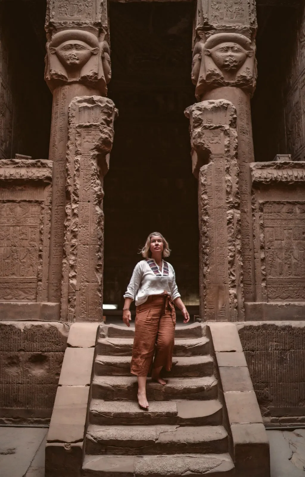 A woman on the steps of a temple in Egypt, the carved Hathor columns of the gateway rising above her