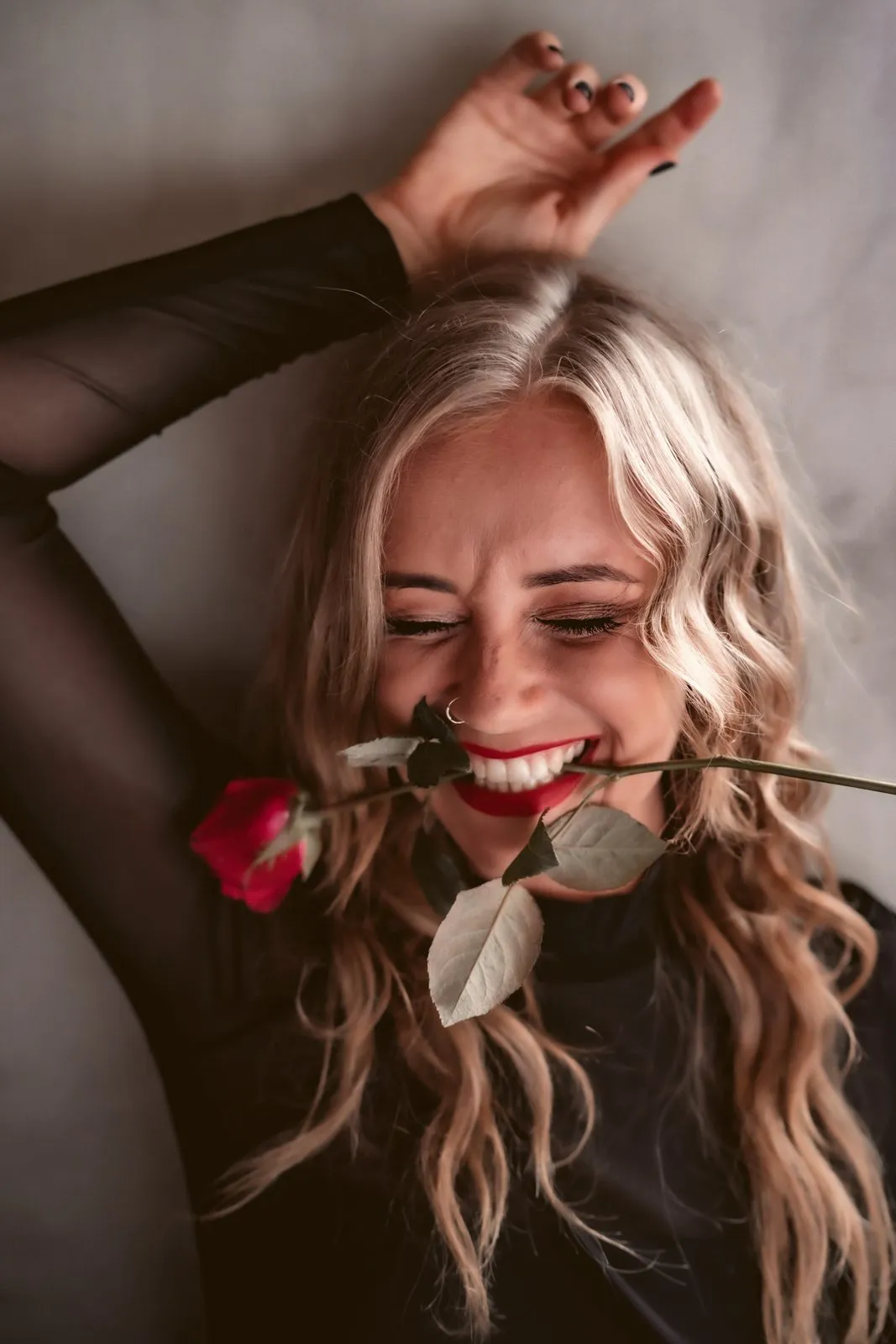 A laughing woman with long blonde hair holding a red rose between her teeth