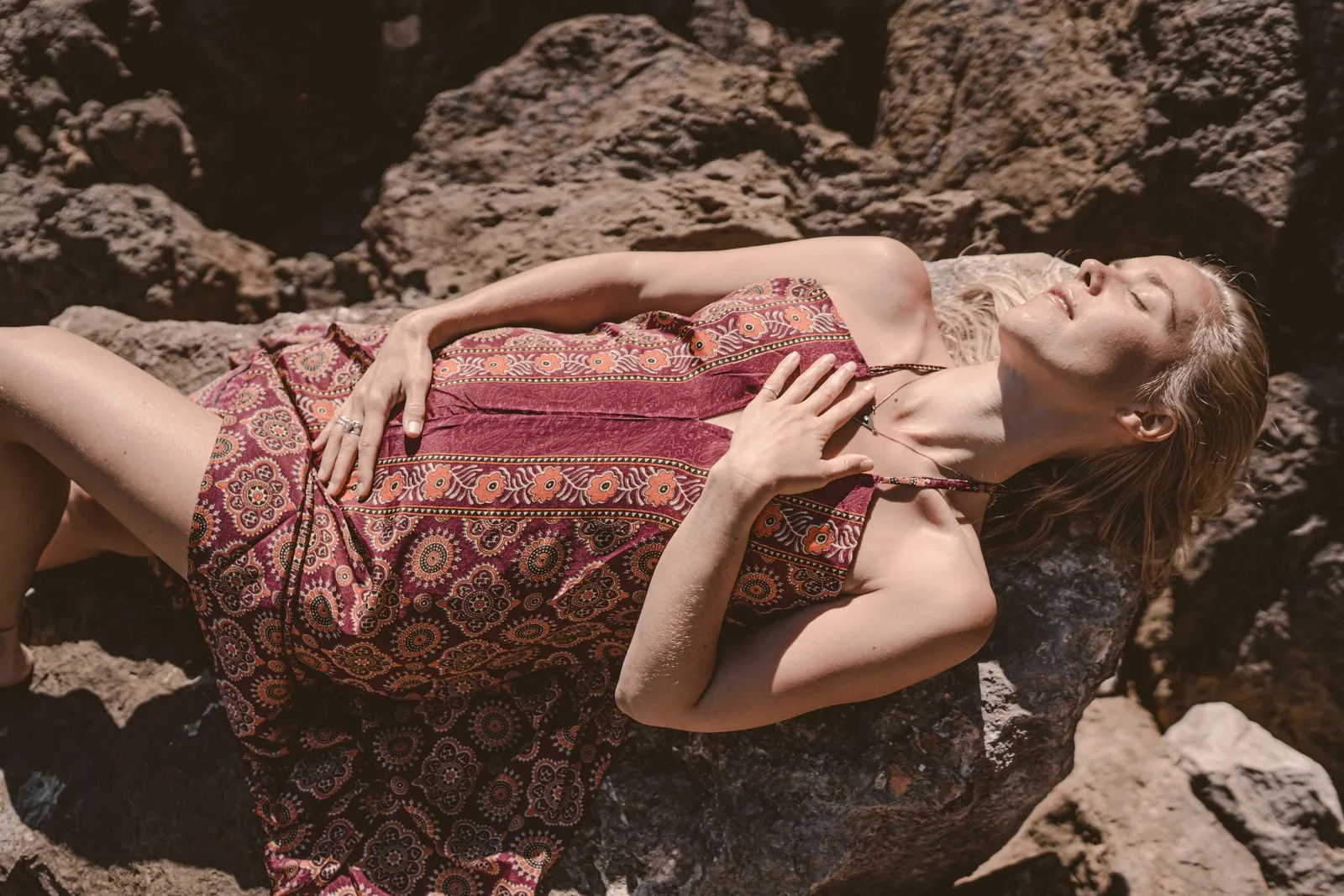 A woman in a patterned dress lying back across the rocks in the sun, a hand on her chest