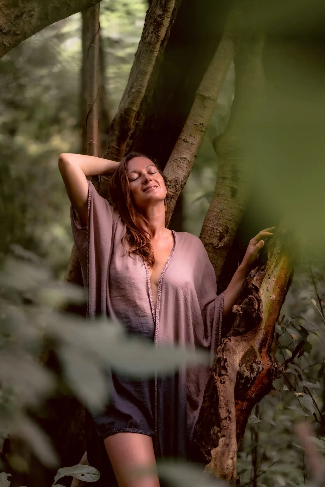A woman in a mauve dress leaning into the roots of a jungle tree, eyes closed