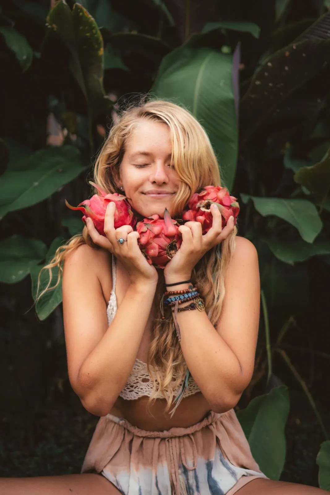 A woman under a banana leaf holding an armful of dragon fruit to her chest, eyes closed and smiling