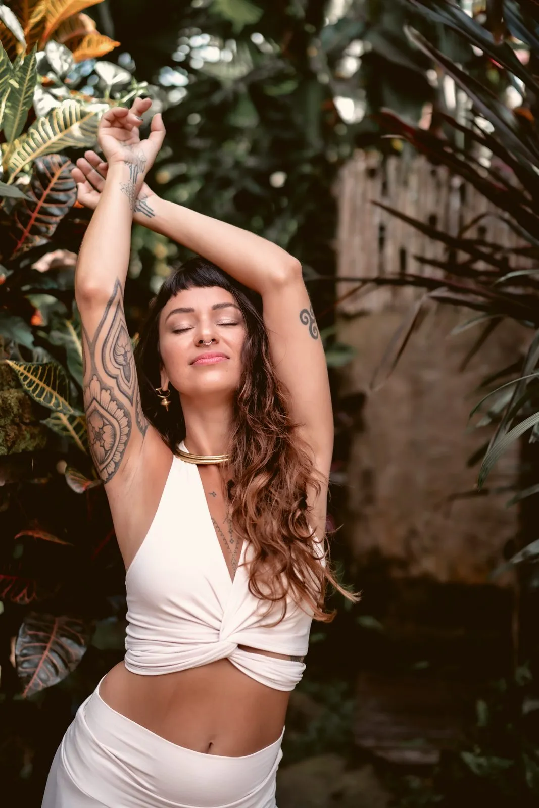 A woman in a white crop top with an arm raised among palm fronds, tattooed and looking down