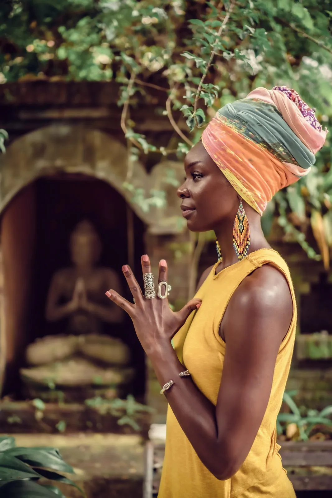 A woman in a yellow dress and a bright head wrap with her hands in prayer before a stone Buddha