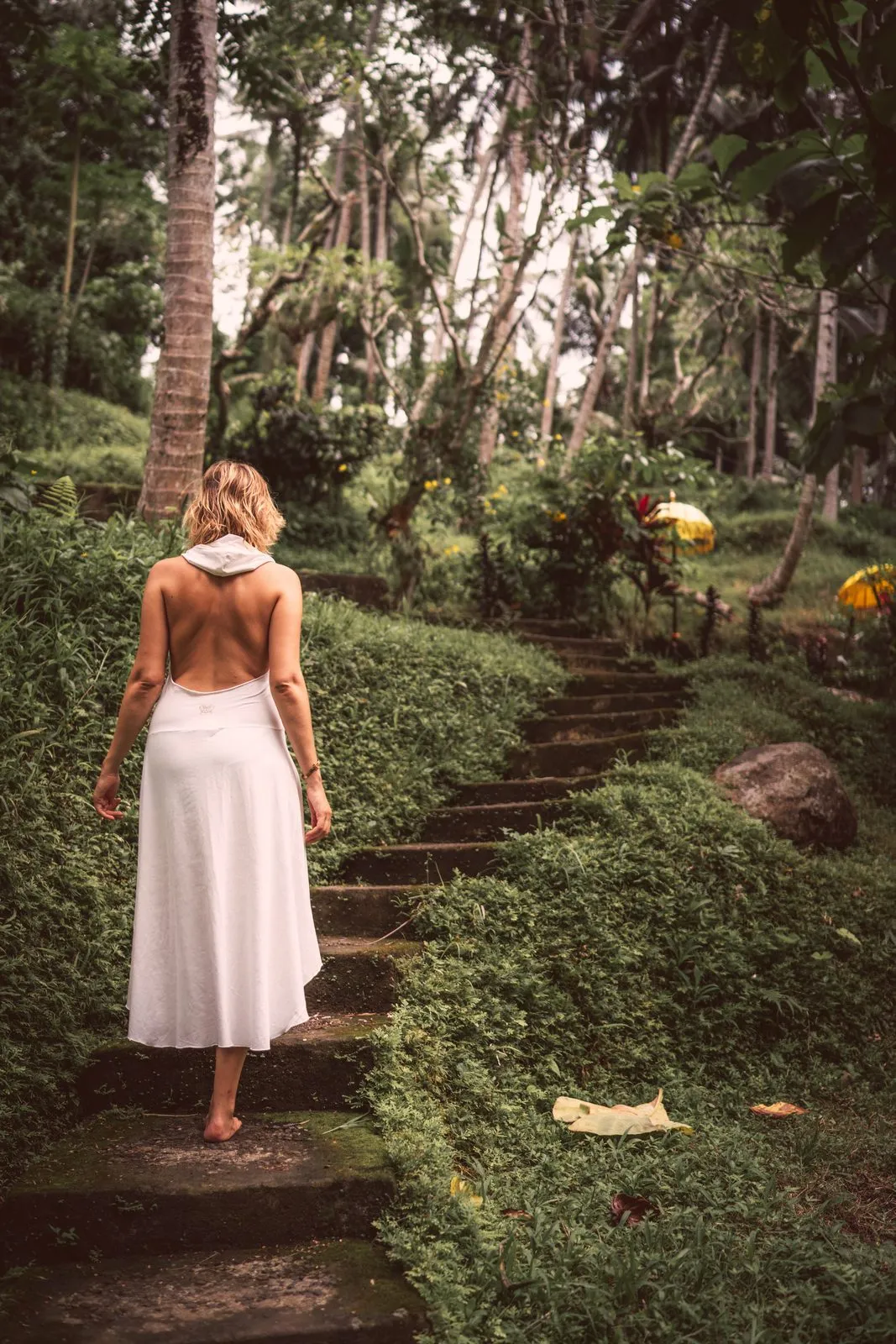 A woman in a white backless dress walking up mossy stone steps through a garden of palms