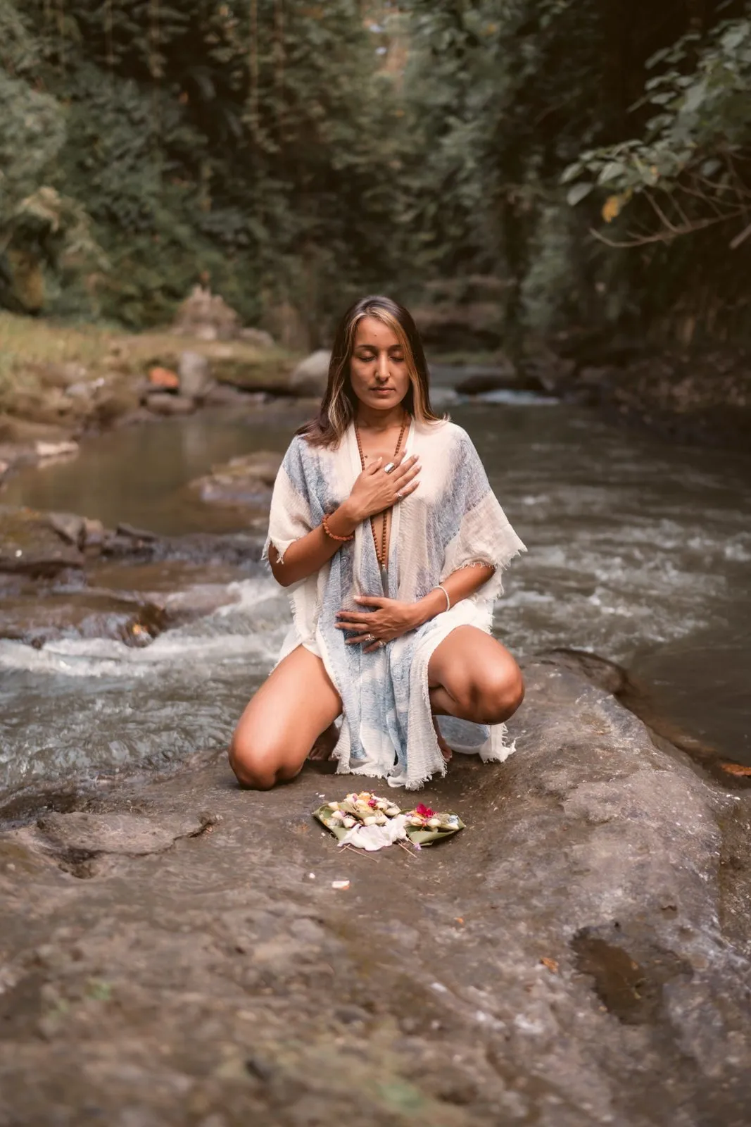 A woman kneeling on a rock in a river with her hands on her heart, offerings laid before her