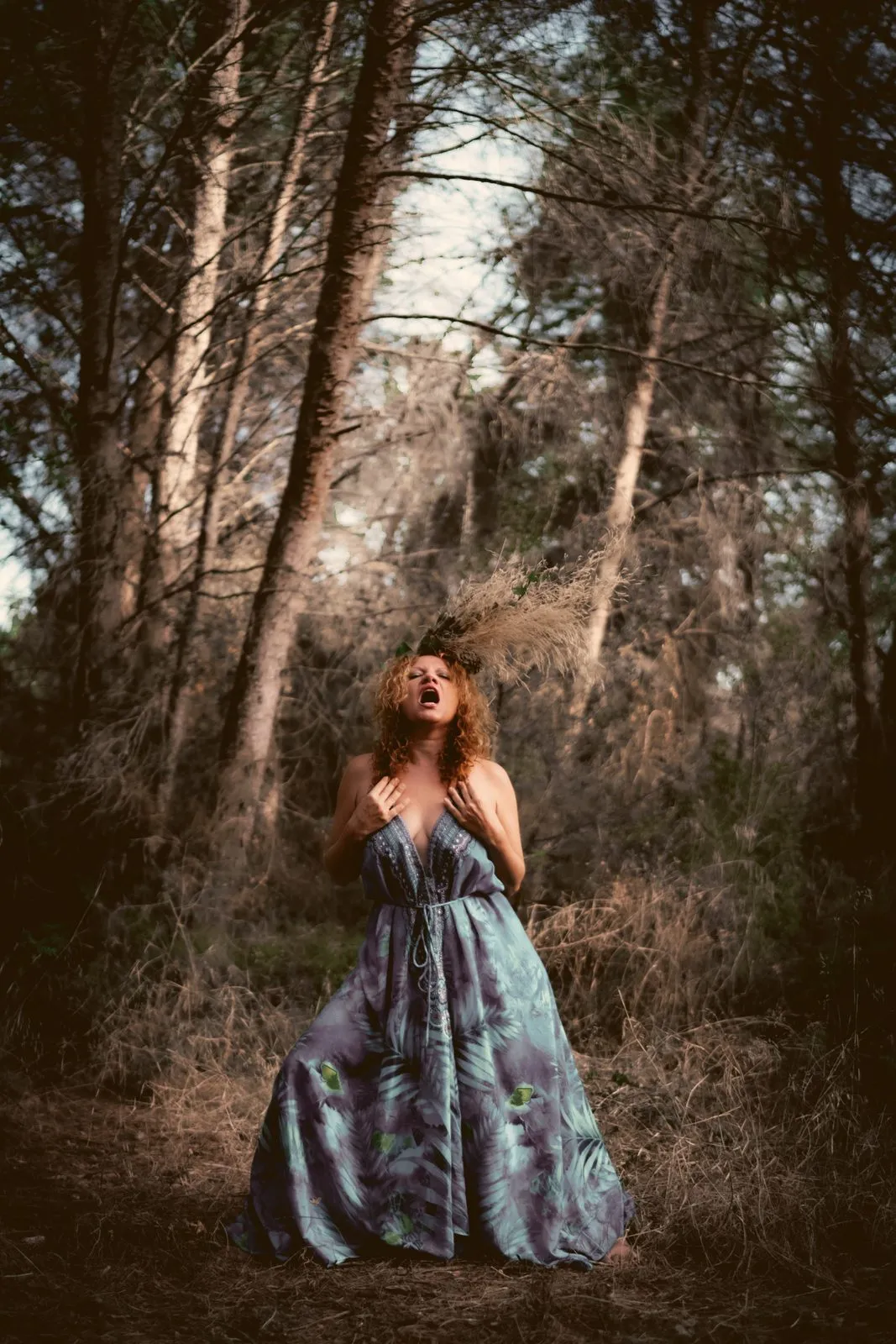 A woman in a long blue and green dress among pine trees, head thrown back