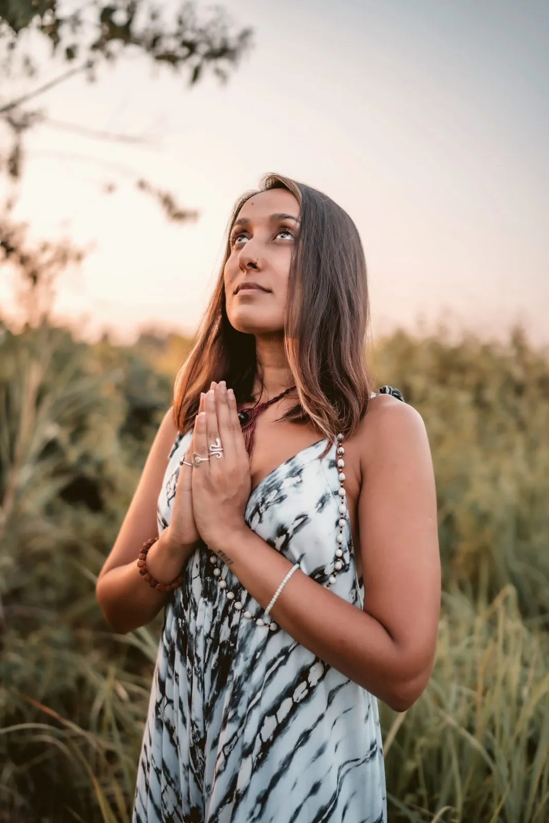 A woman in a tie-dyed dress standing in tall grass with her hands in prayer, looking up