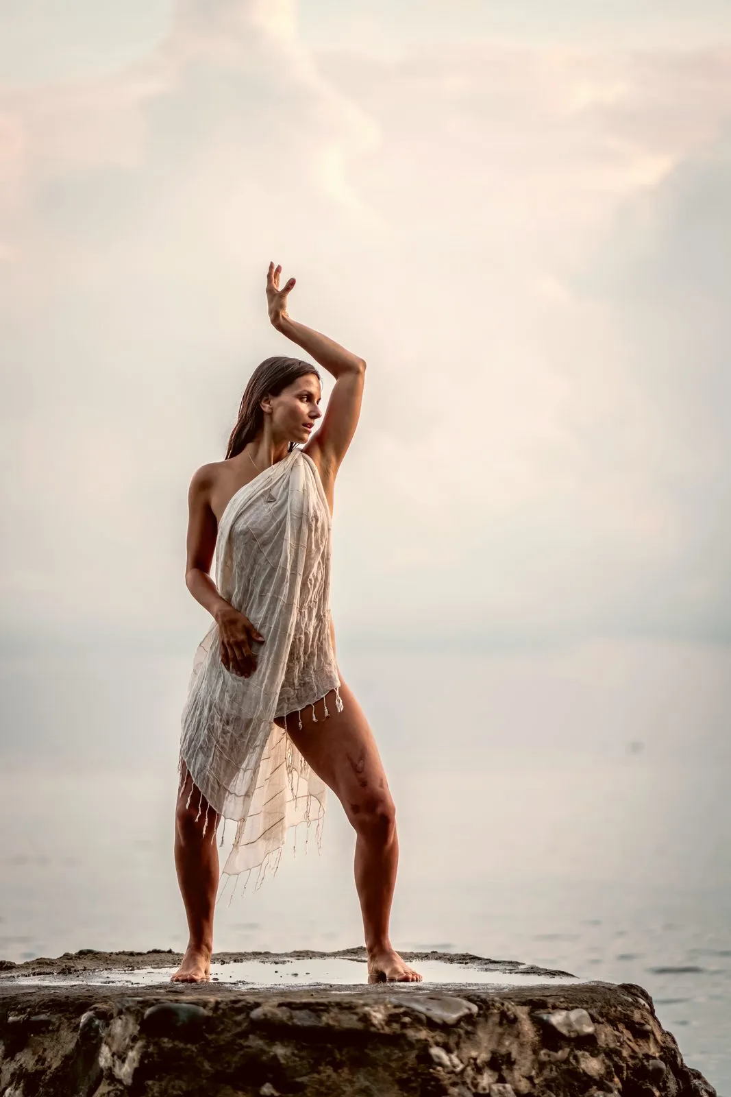 A woman in white standing on a rock at the edge of the sea, one arm curved over her head