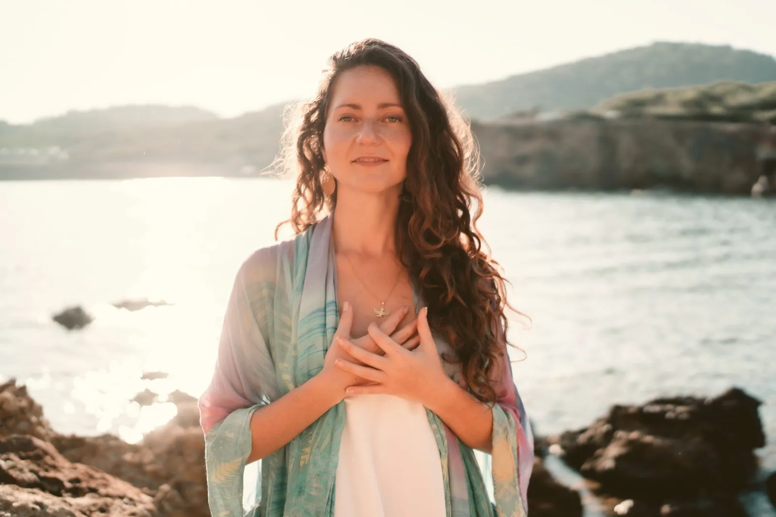 A woman in a pastel kimono with her hands on her heart on a rocky shore, the cove behind her