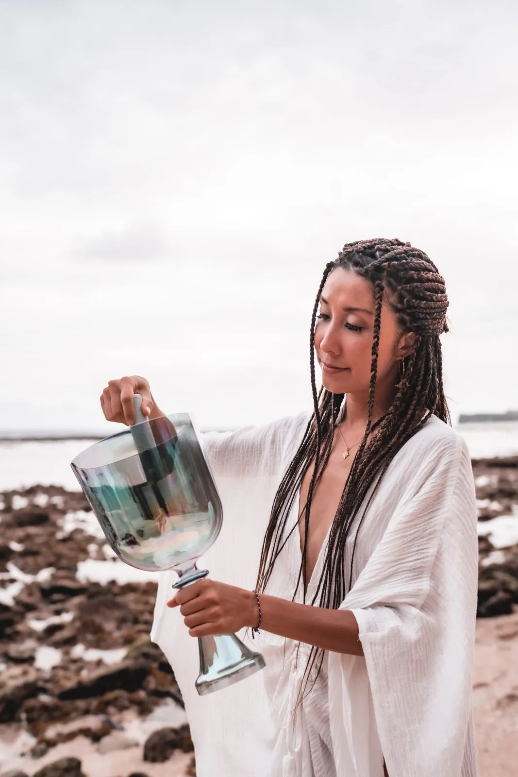 A woman in white with braided hair playing a crystal singing bowl on the rocks by the sea