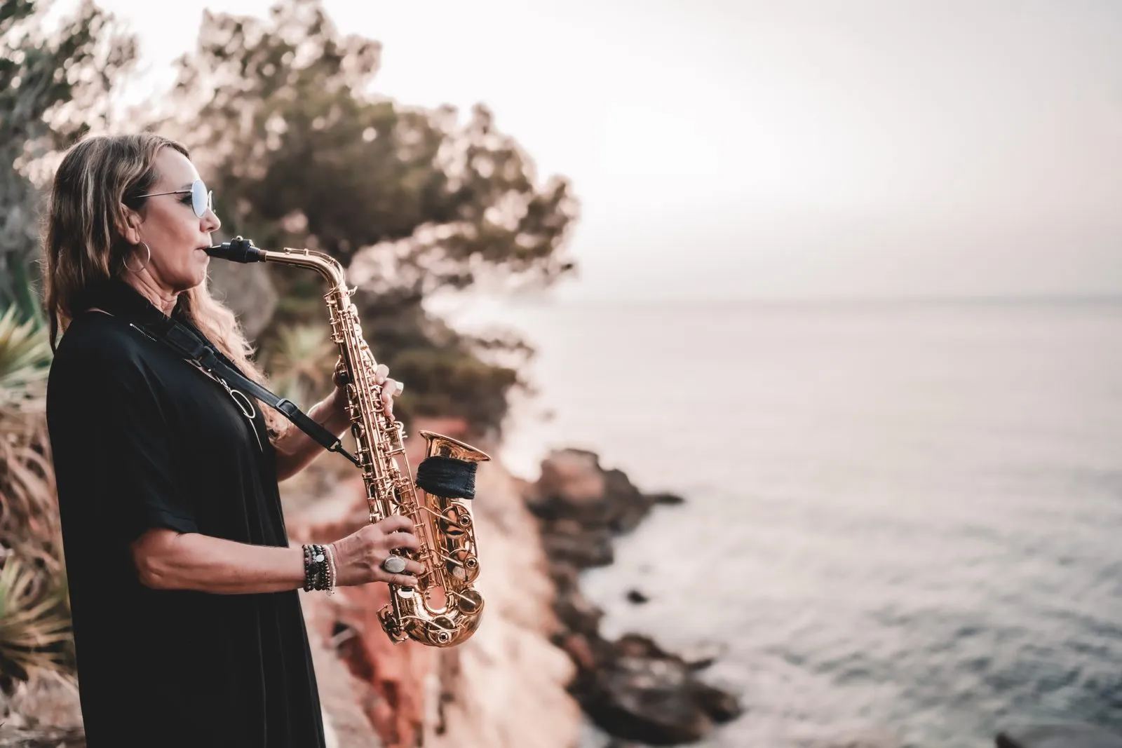 A saxophonist in black playing on a cliff above the sea at dusk