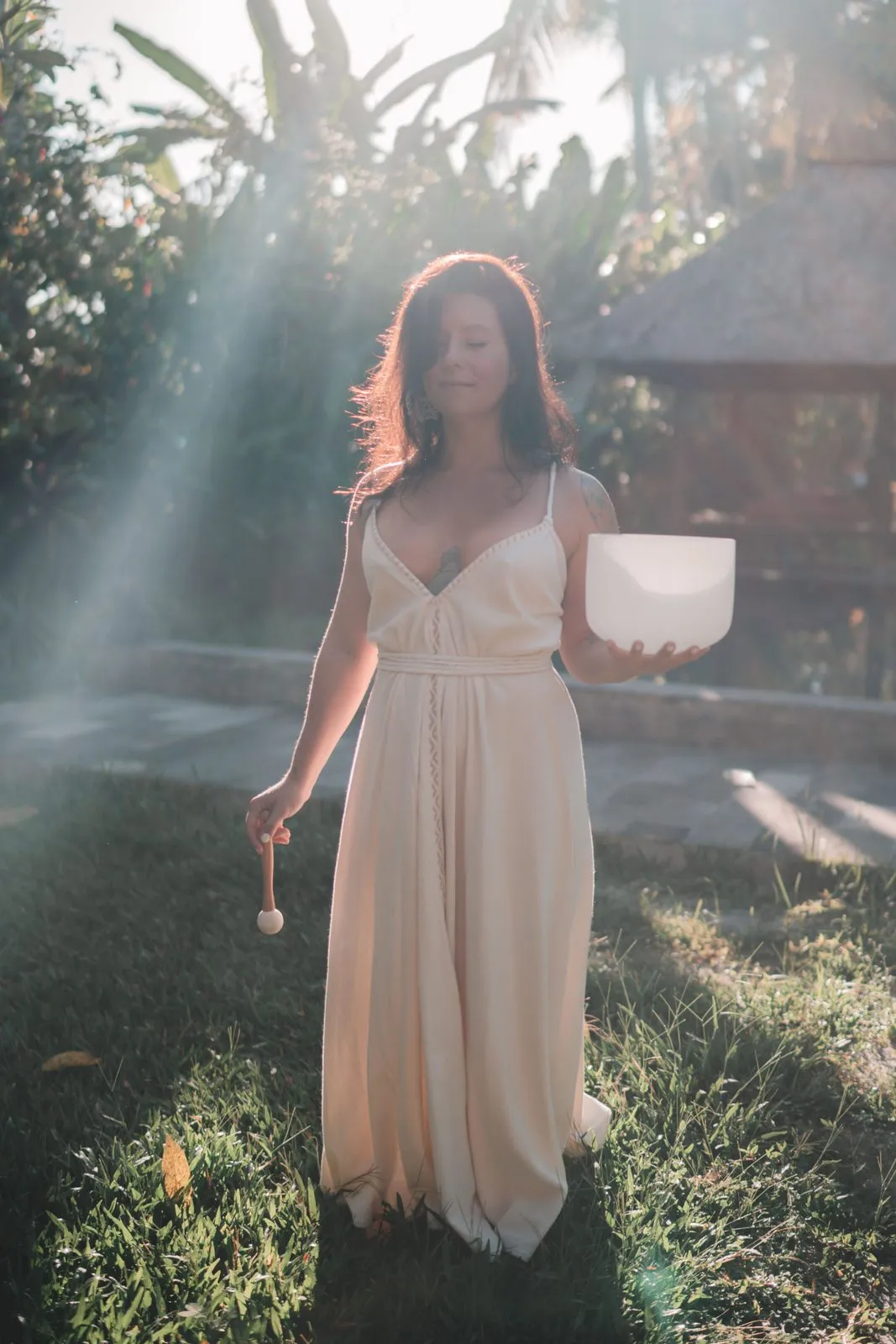 A woman in a cream dress holding a crystal singing bowl in a garden, sunbeams through the trees