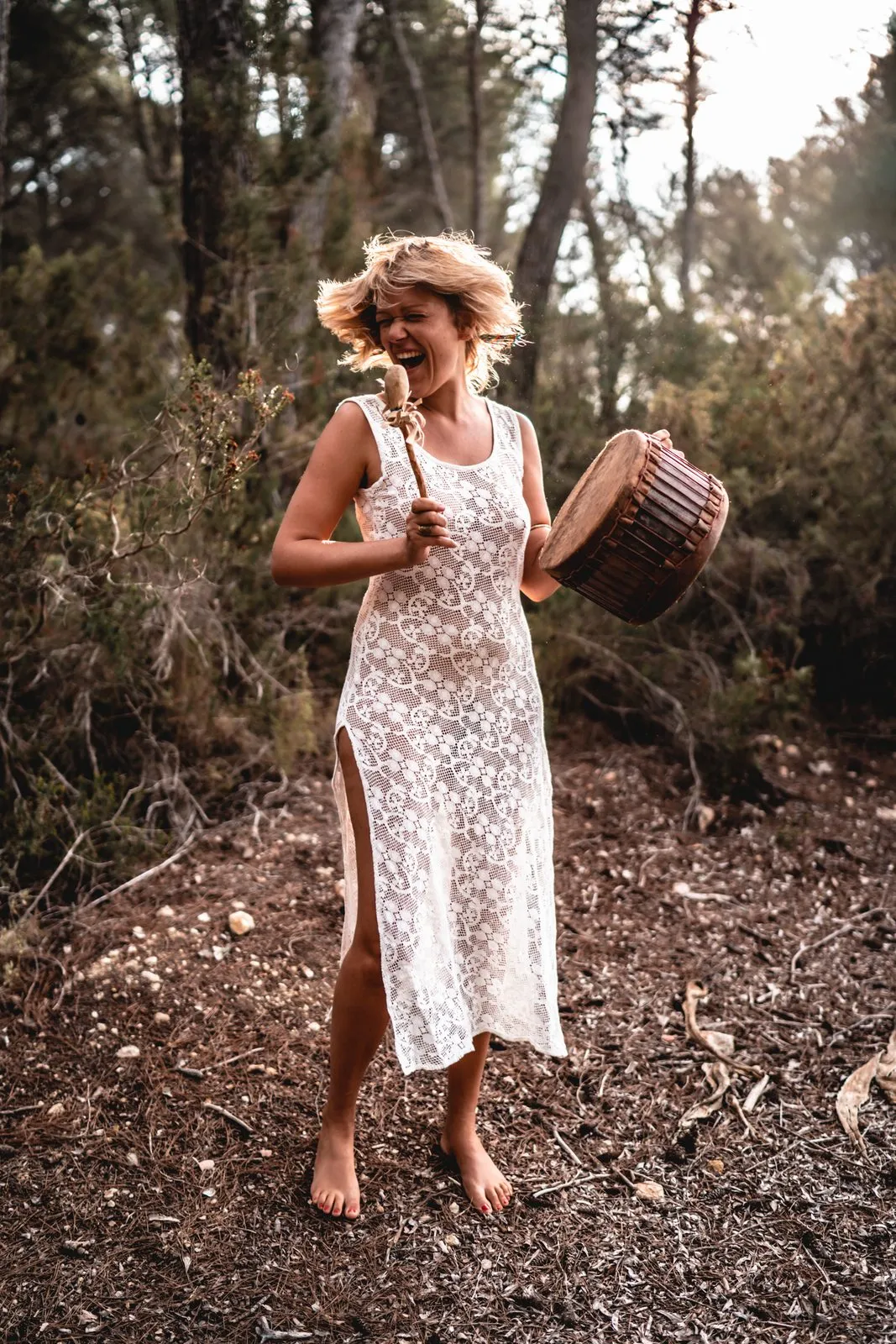 A woman in a white lace dress playing a frame drum barefoot in a pine forest, laughing