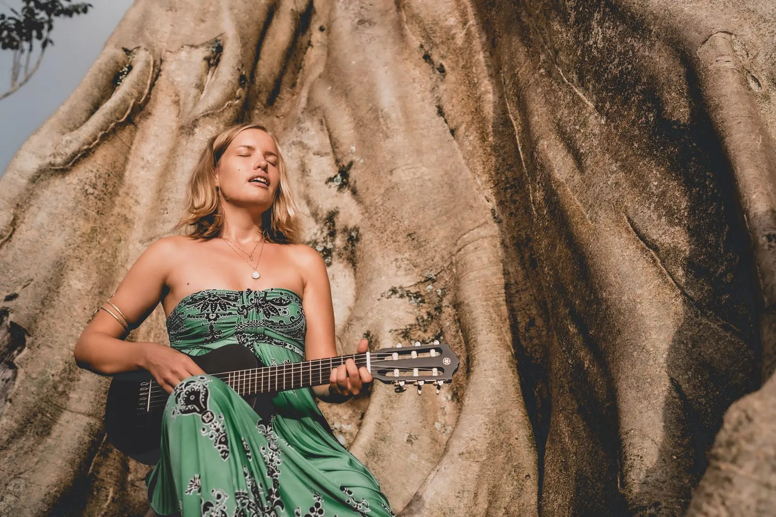 A woman in a green dress playing guitar with her eyes closed, sitting among the roots of a giant tree