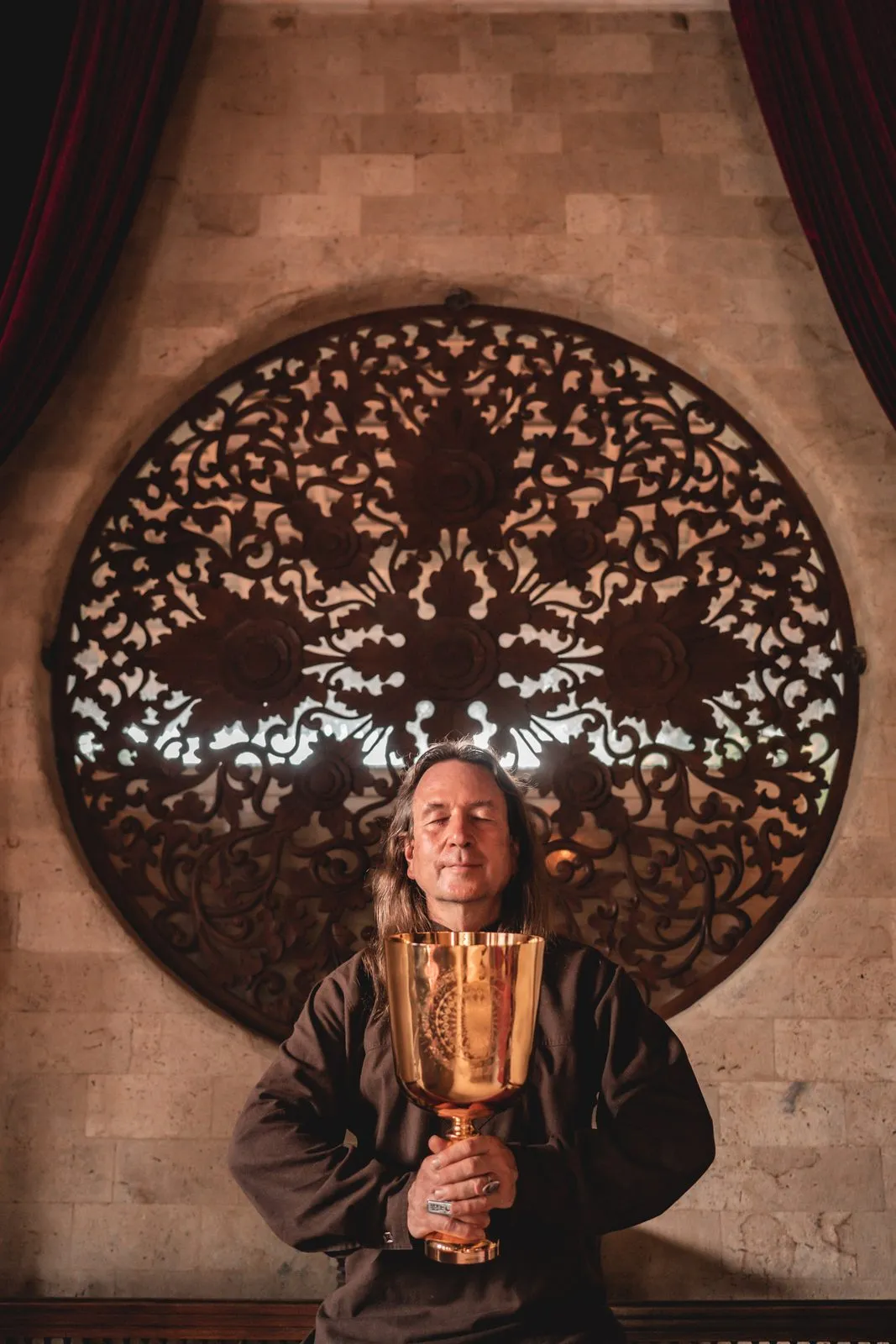 A musician in a brown robe holding a golden chalice beneath a carved wooden mandala window
