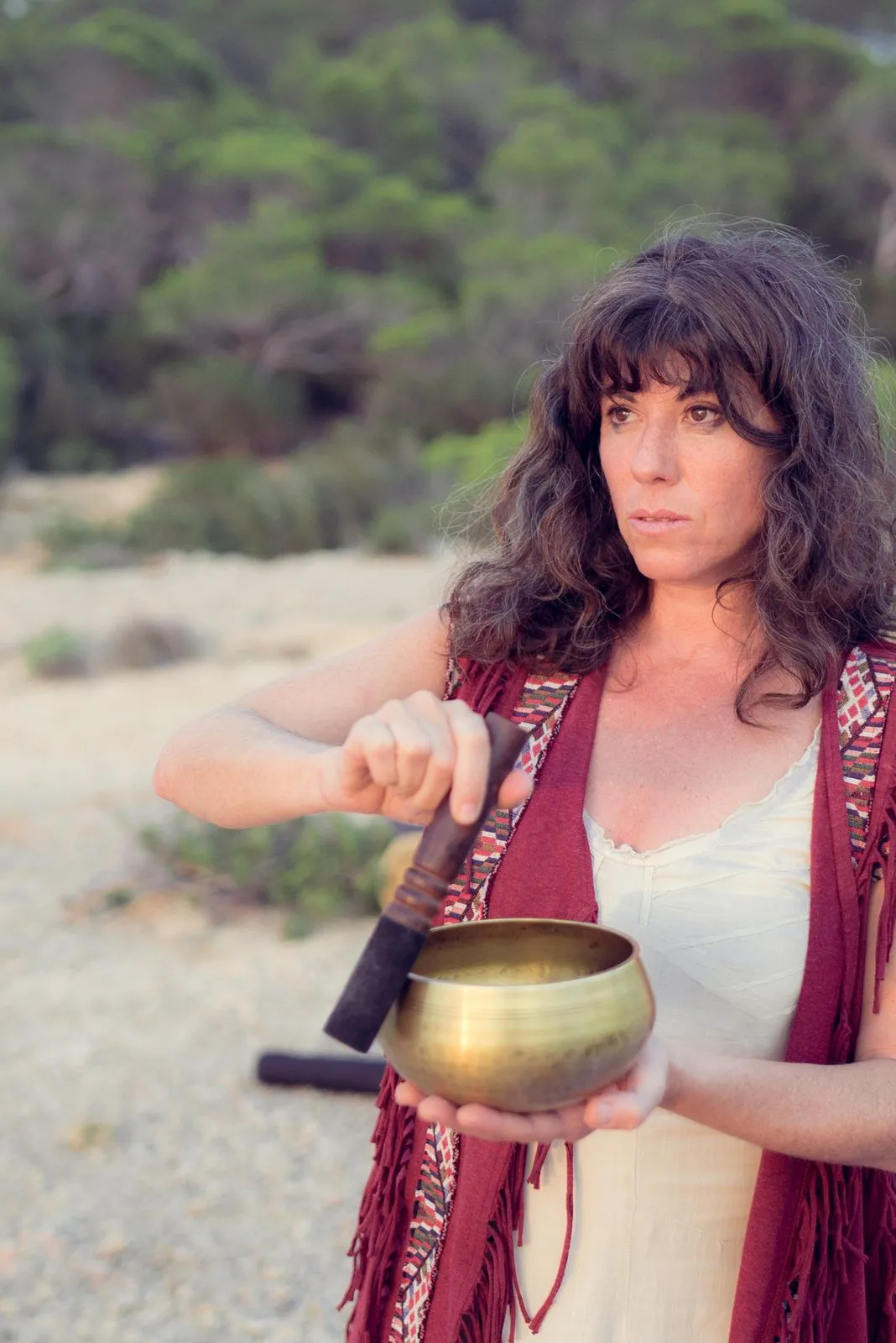 A woman in a red shawl striking a brass singing bowl, pine trees behind her