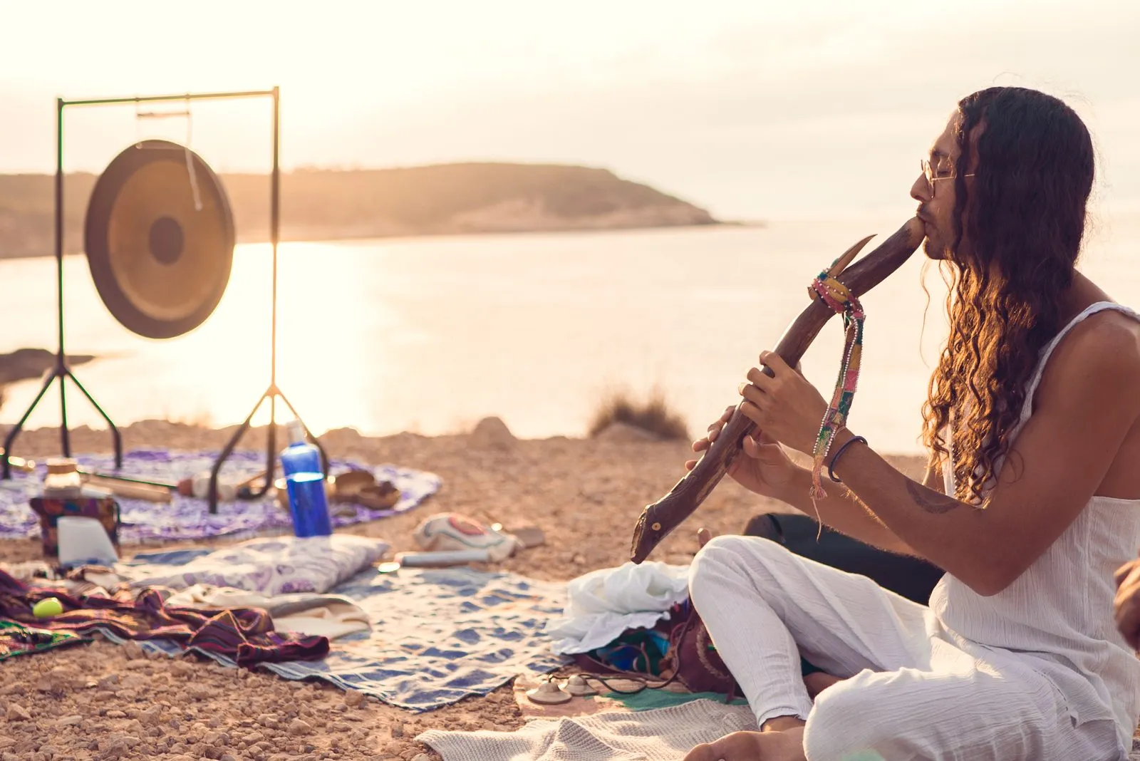 A sound healer playing a wooden flute on the beach at sunrise, a gong on its stand beside her, Ibiza