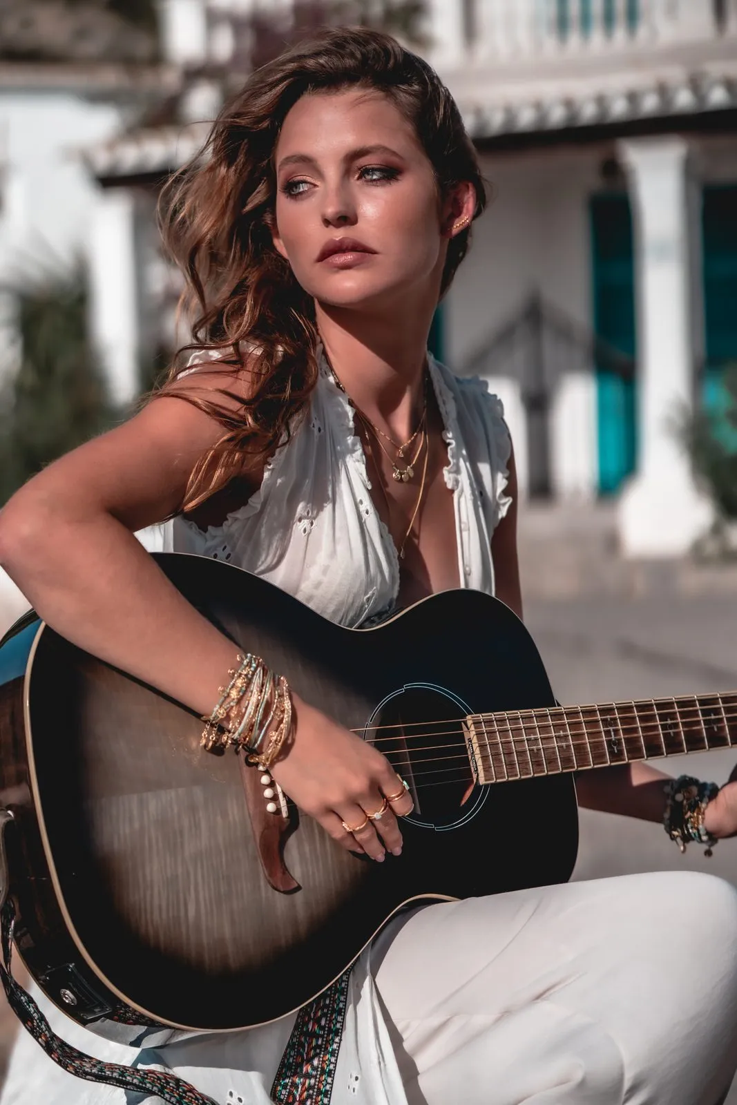 A woman in white with gold bracelets playing a black acoustic guitar, a white villa behind her