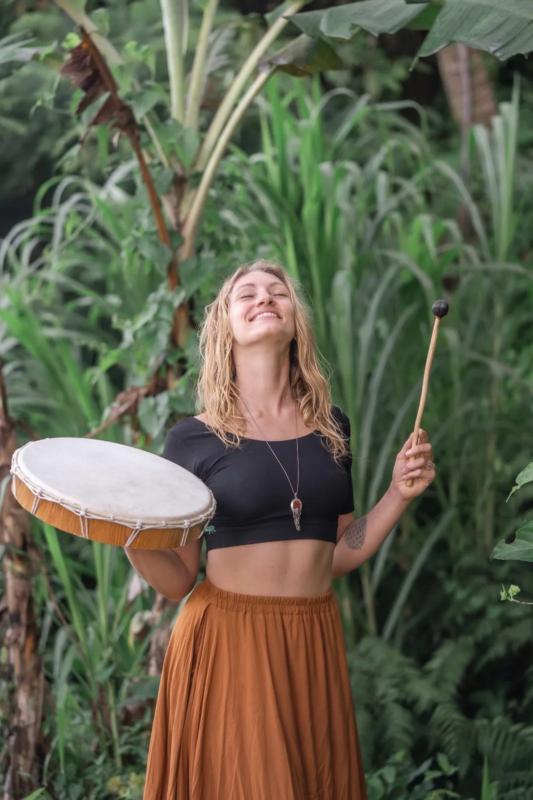 A woman in a rust skirt playing a frame drum among banana leaves, eyes closed