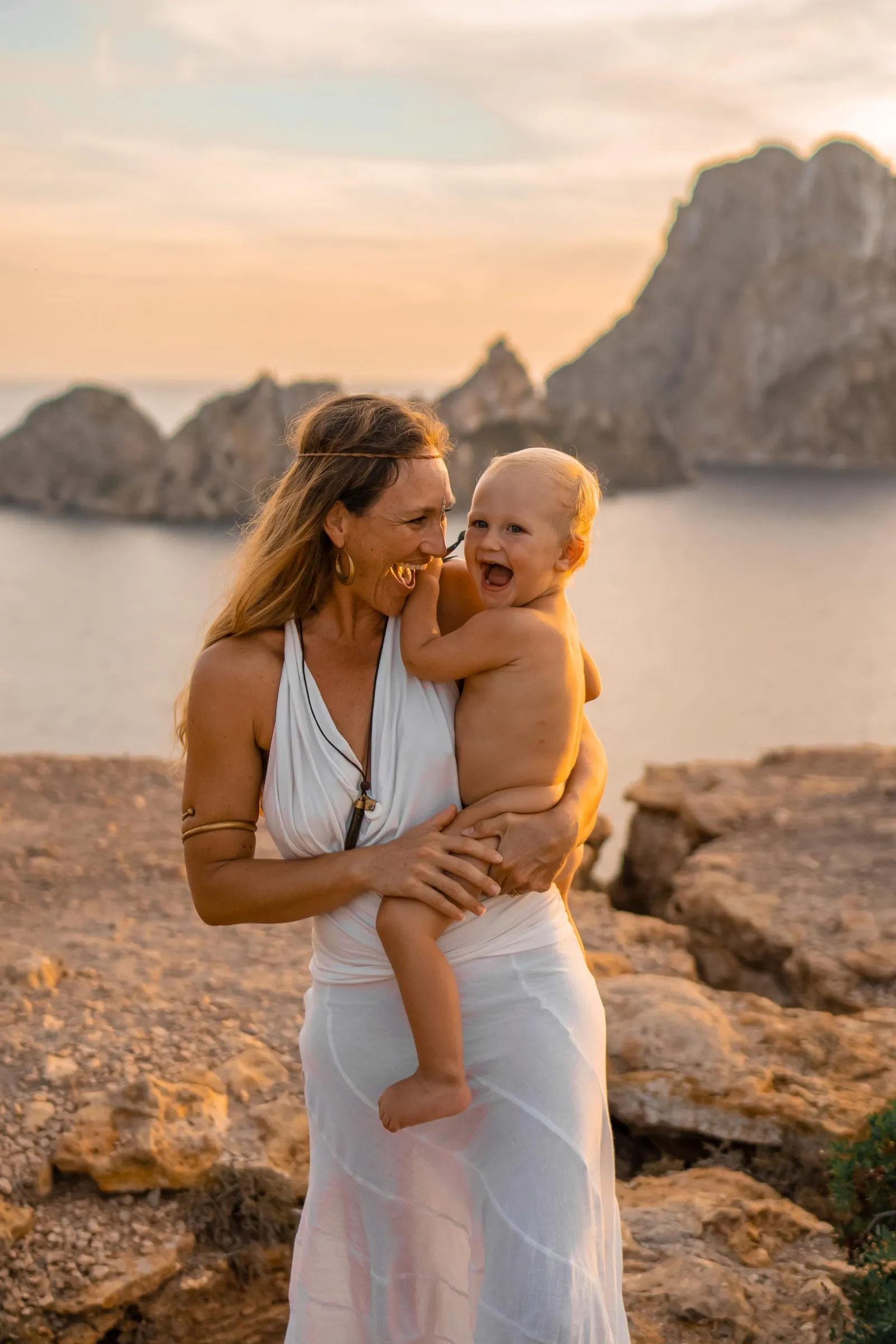 A mother in white holding her laughing toddler on the rocks at sunset, Es Vedrà rising from the sea behind them, Ibiza