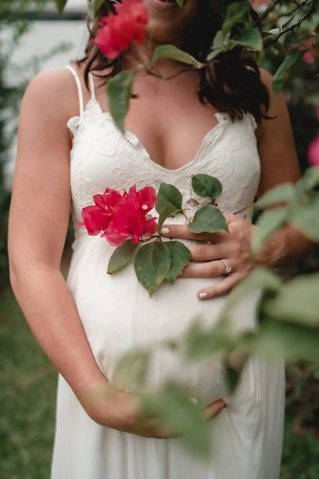 A pregnant woman in white lace holding a sprig of red bougainvillea to her chest