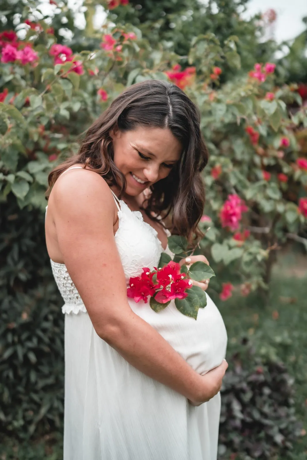 A pregnant woman in a white dress smiling down at her belly among red bougainvillea