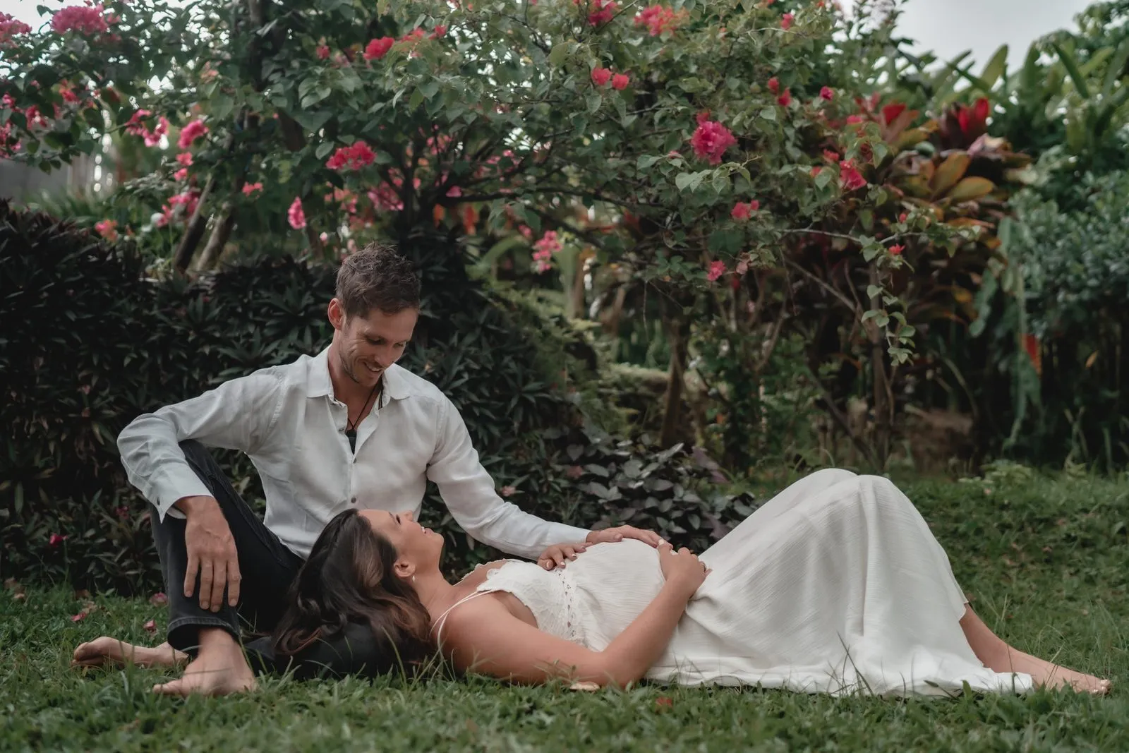 An expectant couple lying together on the grass beneath a wall of red bougainvillea