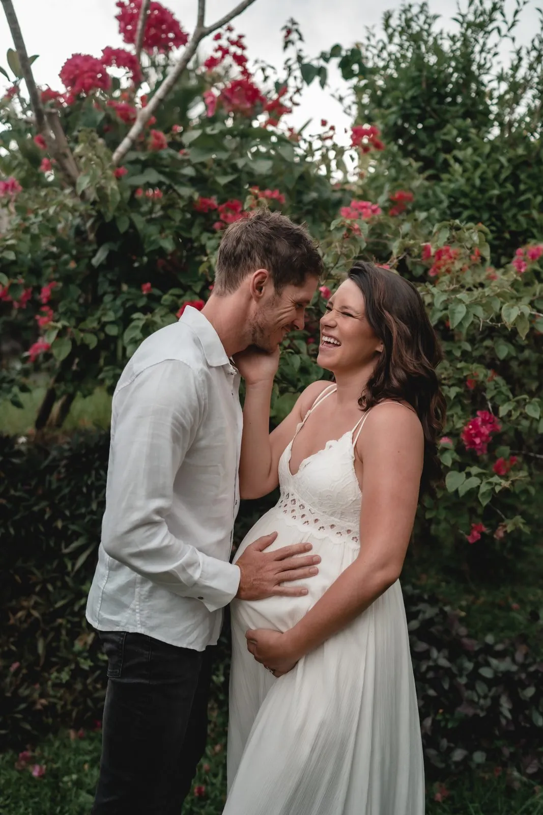 An expectant couple in a garden of red flowers, his hands on her belly, laughing together