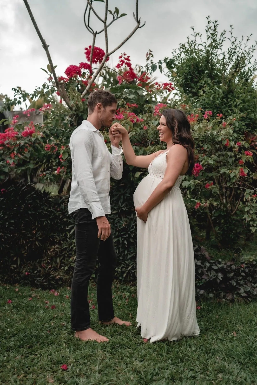 A man kissing the hand of his pregnant partner in a white dress, red bougainvillea behind them