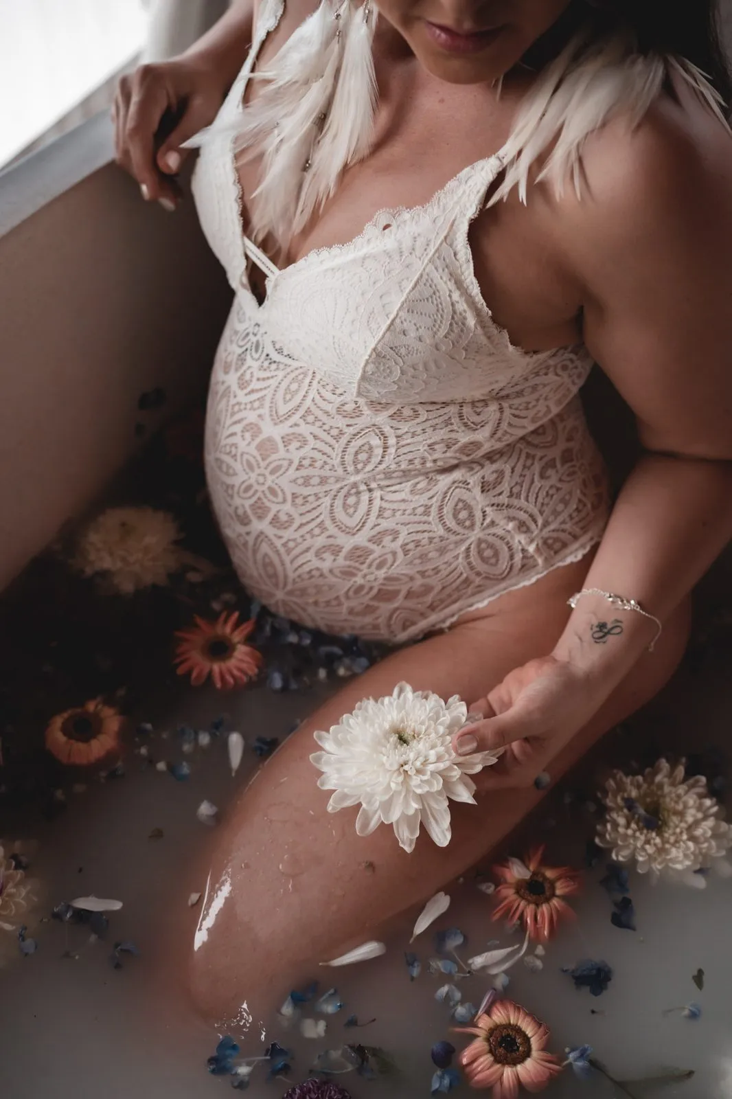 A pregnant belly in white lace in a milk bath, a white dahlia in her hand and dried flowers floating