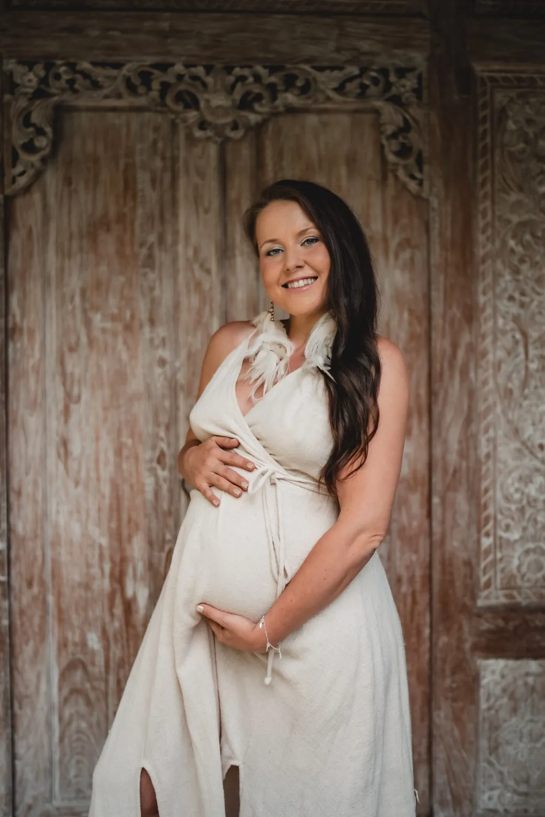 A pregnant woman in a white wrap dress cradling her belly before a carved wooden door