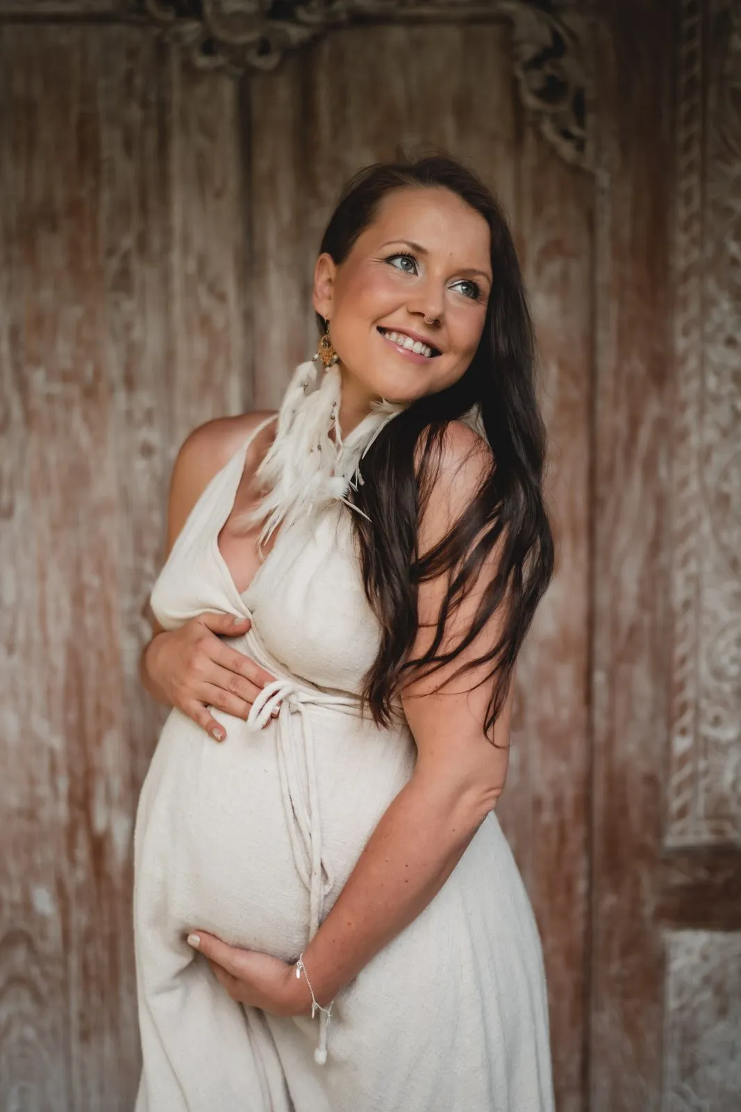 A smiling pregnant woman in a white halter dress with both hands on her belly, an old wooden door behind
