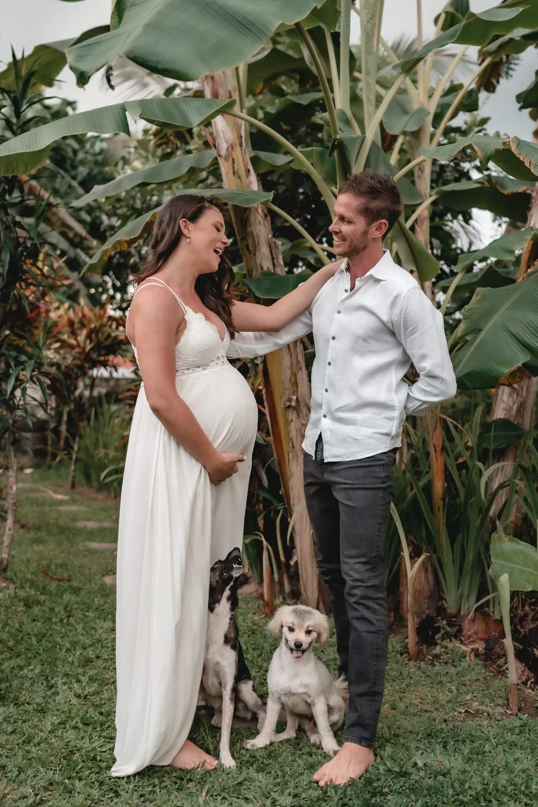 An expectant couple in a Balinese garden with their dog sitting at their feet, she in white