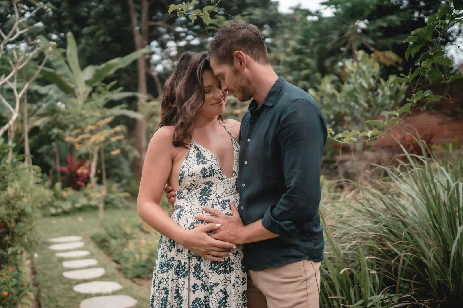 An expectant couple forehead to forehead on a garden path, his hand on her belly, Bali
