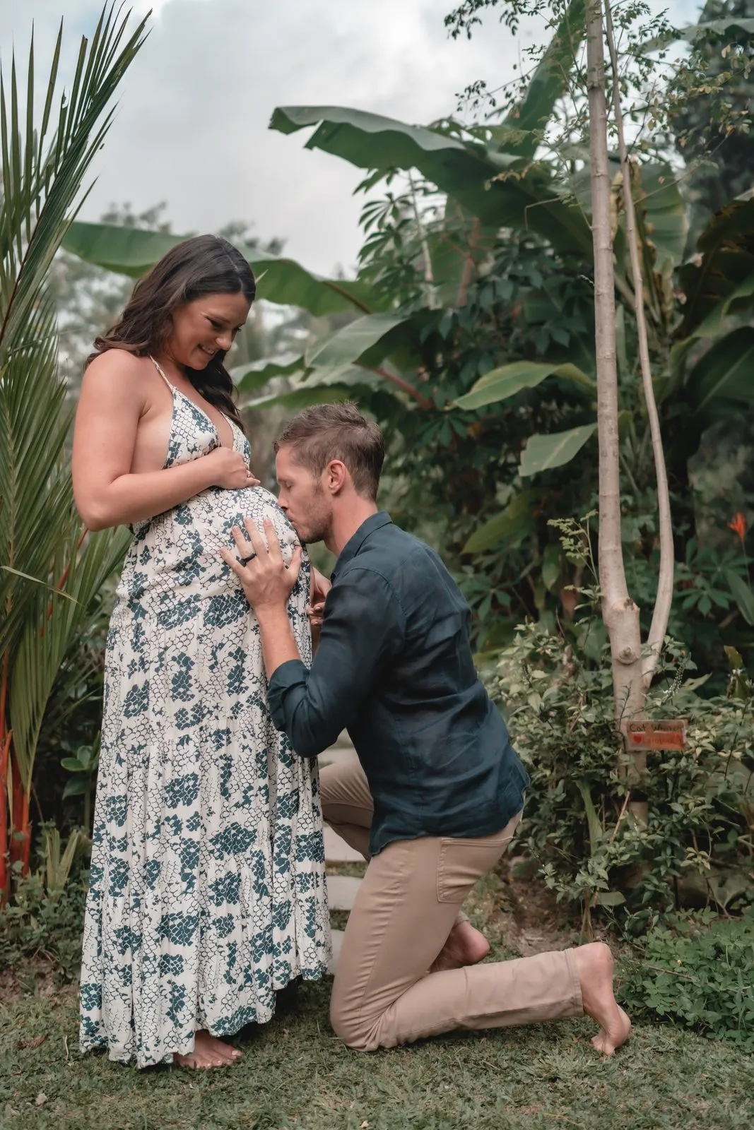 A man kneeling to kiss his pregnant partner's belly beneath banana palms in a garden